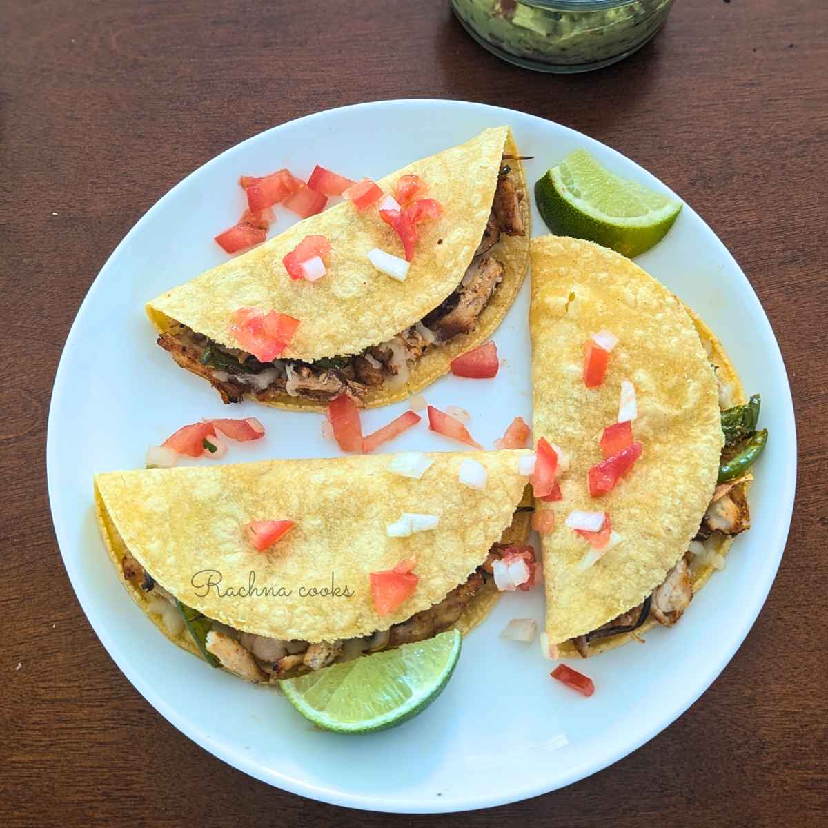 3 chicken tacos served on a white plate with chopped onion, tomatoes and slices of lemon.