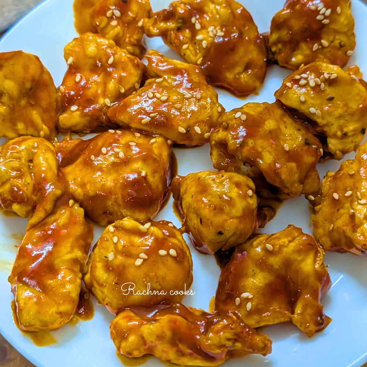 Close up of glossy gochujang chicken bites on a white plate with a garnish of sesame seeds.