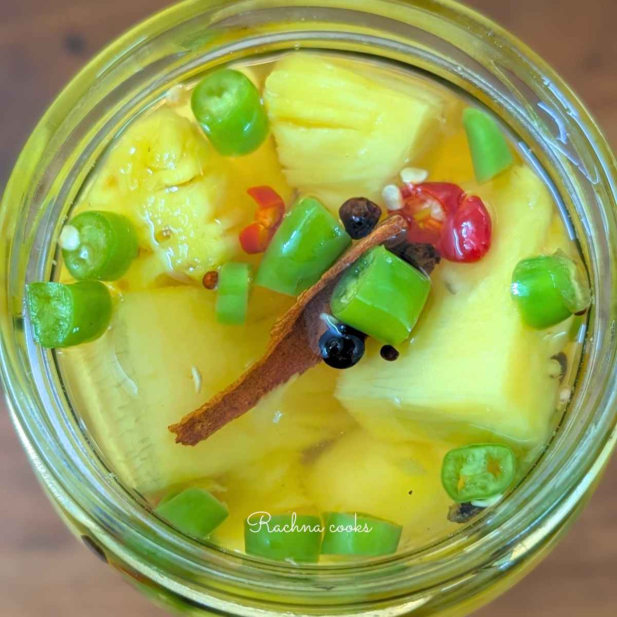 Top view of pickled pineapple chunks in a glass jar with green and red sliced chillies, black peppercorn,mustard seeds and cinnamon floating in pickling liquid.