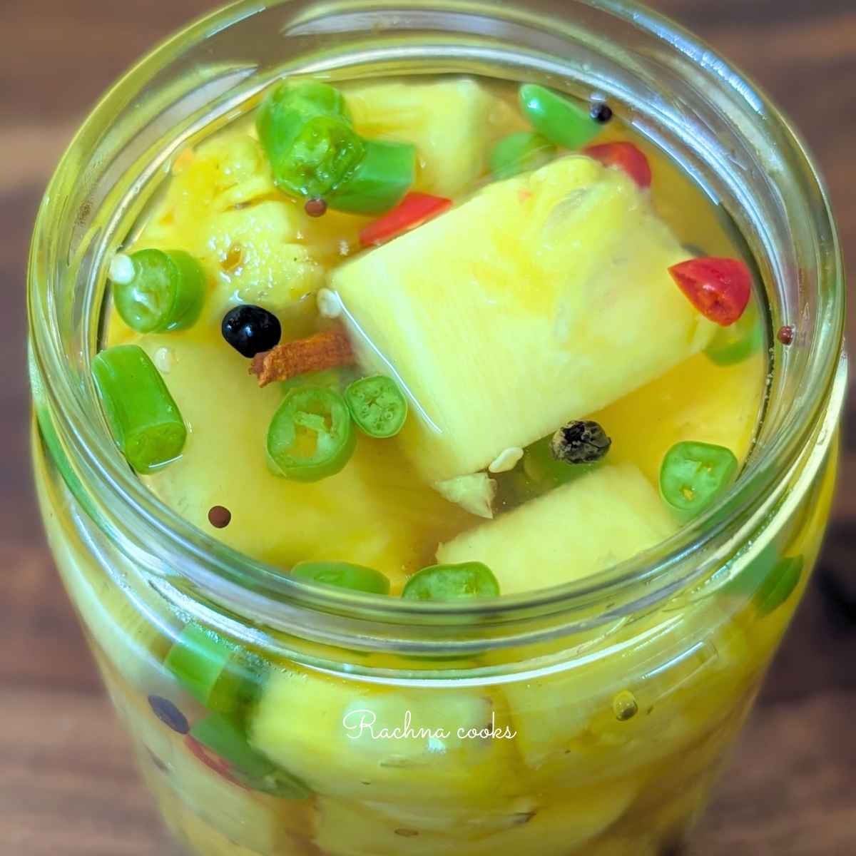 Side view of glass jar of pickled pineapple chunks in a glass jar with green and red sliced chillies, black peppercorn,mustard seeds and cinnamon floating in pickling liquid.