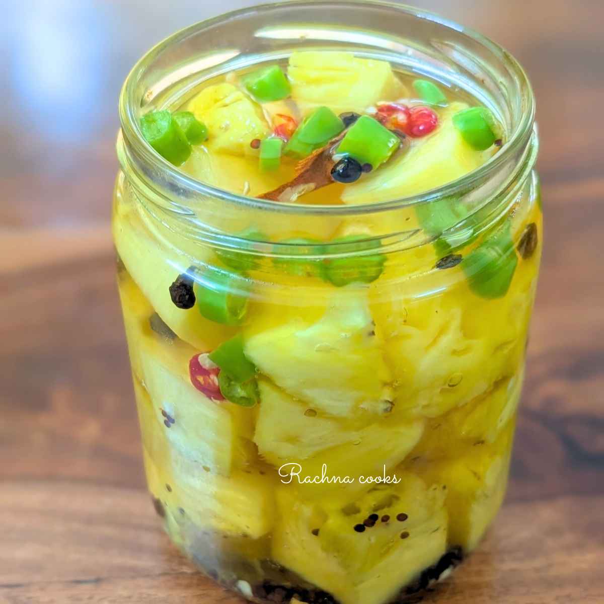 A tall glass jar of pickled pineapple chunks in a glass jar with green and red sliced chillies, black peppercorn,mustard seeds and cinnamon floating in pickling liquid.