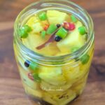 Side view of pickled pineapple chunks in a glass jar with green and red sliced chillies, black peppercorn,mustard seeds and cinnamon floating in pickling liquid.