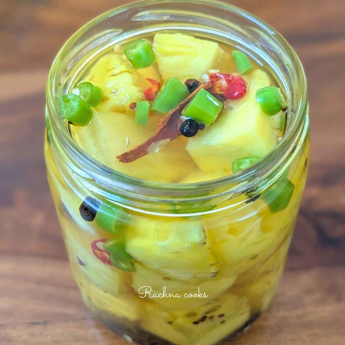Side view of pickled pineapple chunks in a glass jar with green and red sliced chillies, black peppercorn,mustard seeds and cinnamon floating in pickling liquid.